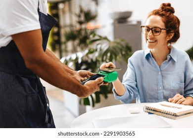 Woman making a cashless payment at a cafe using the NFC technology on her smartphone. Happy customer doing a simple tap on the terminal to pay her bill quickly and securely without cash or card. - Powered by Shutterstock