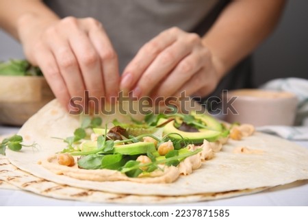 Woman making delicious hummus wrap with vegetables at table, closeup