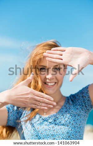 Woman making a finger frame with her hands framing her face as she stands against a sunny blue sky