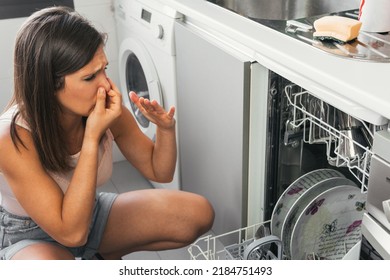 Woman making gestures of bad smell when opening her automatic dishwasher. Young girl with a face of disgust and holding her nose at the malfunction of one of her appliances.