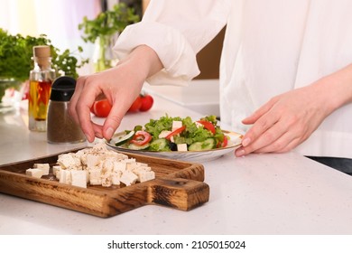 Woman Making Greek Salad With Cut Feta Cheese On Table In Kitchen