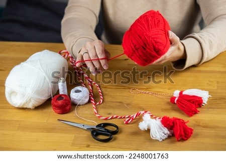 Woman making handmade traditional martisor, from red and white strings with tassel. Symbol of holiday 1 March, Martenitsa, Baba Marta, beginning of spring in Romania, Bulgaria, Moldova