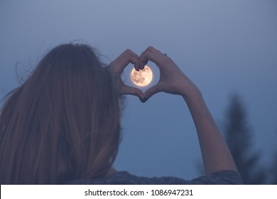 Woman making heart shape with her hands over full moon.