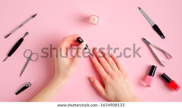 Woman making manicure herself. Female\
hands with nail polish and manicure tools on pink background, view\
from above. Self-care beauty treatment\
concept