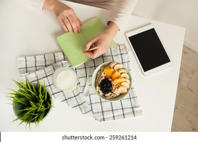 Woman Making Notes In Notepad With Porridge, Milk And Tablet On Table