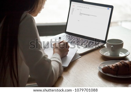 Woman making notes reading email letter on laptop screen sitting at cafe table, young lady working with computer mailing clients writing information in personal organizer, close up over shoulder view