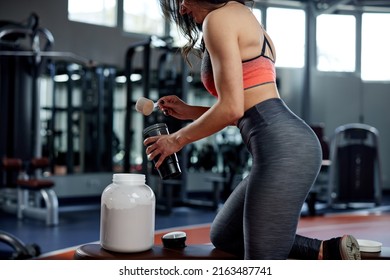 A Woman Is Making A Protein Shake After A Workout In The Gym. She Uses A Measuring Cup To Mix The Protein Powder In Her Cup.