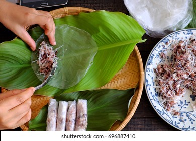 Woman making spring rolls or cha gio at home, homemade food stuffing from meat and wrapper by rice paper, hand rolling Vietnamese egg roll on green leaf background