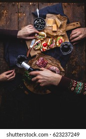 A Woman And A Man Eat A Set Of Bruschetta, Vegetables, Arugula, Dried Cured Sausage, A Corkscrew On An Old Wooden Table. Mediterranean Snacks. People Are Drinking Wine With Italian Food.