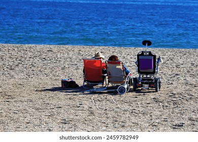 a woman and a man, the family of a disabled person, relax in sun loungers by the sea next to an electric wheelchair - Powered by Shutterstock