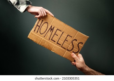A woman and a man hold a cardboard sign with the inscription HOMELESS. Dark background. Hands close-up. The concept of helping vagabonds and panhandle.