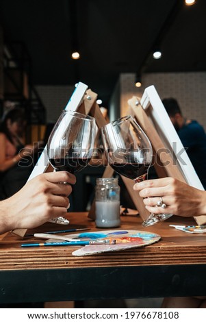 Woman and man making a toast with glasses of wine, next to canvases on wooden easels.