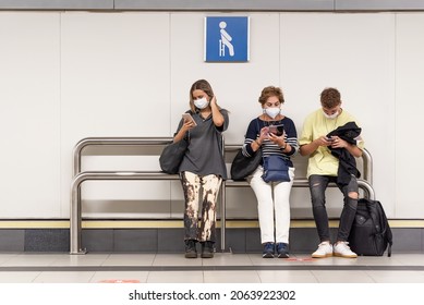 A Woman, A Man And An Older Woman Sitting At A Subway Stop Looking At Their Cell Phones Wearing Mask