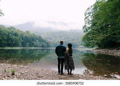 Woman and man stand by the lake in the park and look at the mountains. Back view