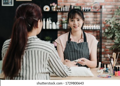 Woman Manicurist Giving Manicure To Unrecognized Asian Female Client At Nail Salon. Confident Young Girl Beautician Face Camera Smiling In Beauty Workshop. Elegant Nail Specialist Worker In Studio.