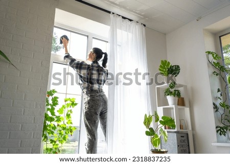 Woman manually washes the window of the house with a rag with spray cleaner and mop inside the interior with white curtains. Restoring order and cleanliness in the spring, cleaning servise
