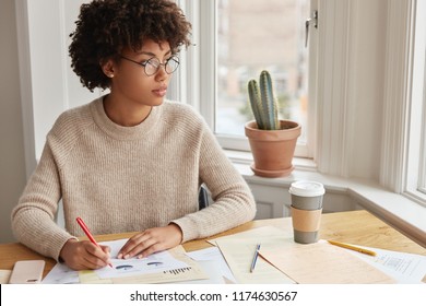 Woman marketing expert writes and makes diagram, thinks about work, wears round glasses and sweater, drinks takeaway coffee. Intelligent dark skinned female businesswoman creats business report