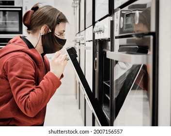 Woman In Mask Against Virus In A Store Chooses For Buy A Kitchen Stove. She Looks Into The Oven And Carefully Examines The Appliance. Concept Of Buying New Gadgets And Household Devices For The Home