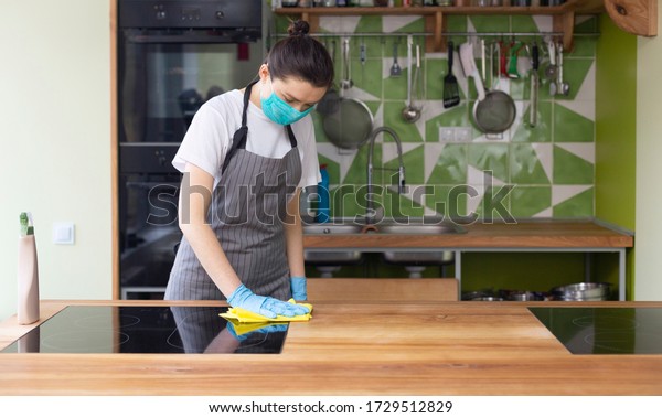 Woman\
in mask and gloves cleaning kitchen electric ceramic hob, polishing\
glass with microfiber cloth, quarantine,\
panorama