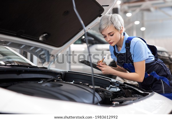 Woman Mechanic writing\
on clipboard at repair garage, wearing uniform overalls. Young\
mechanic engineer female taking notes on clipboard for examining a\
vehicle, side view