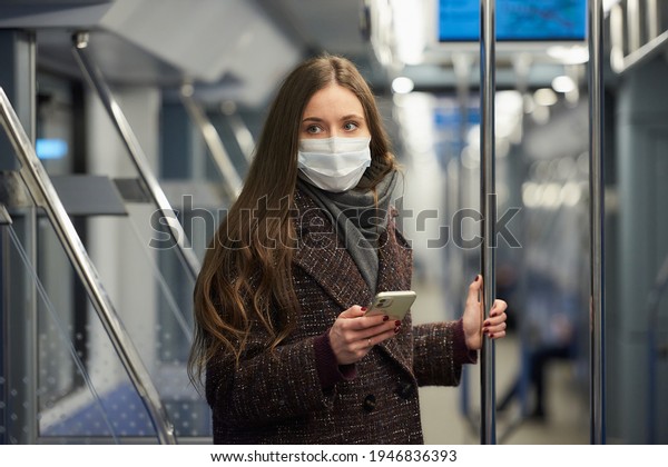 Woman in a medical face mask to avoid the spread of\
coronavirus is standing and staring to the side in a modern subway\
car. A girl in a surgical mask against COVID-19 is taking a ride on\
a metro train