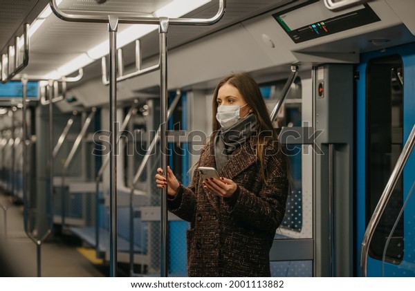 A woman in a medical face mask to avoid the spread\
of coronavirus is standing and holding a phone in a modern subway\
car. A girl in a surgical mask against COVID-19 is taking a ride on\
a metro train.