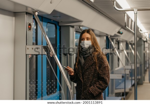 A woman in a medical face mask to avoid the spread\
of coronavirus is standing near doors and staring to the side in an\
empty subway car. A girl in a surgical mask is taking a ride on a\
metro train.