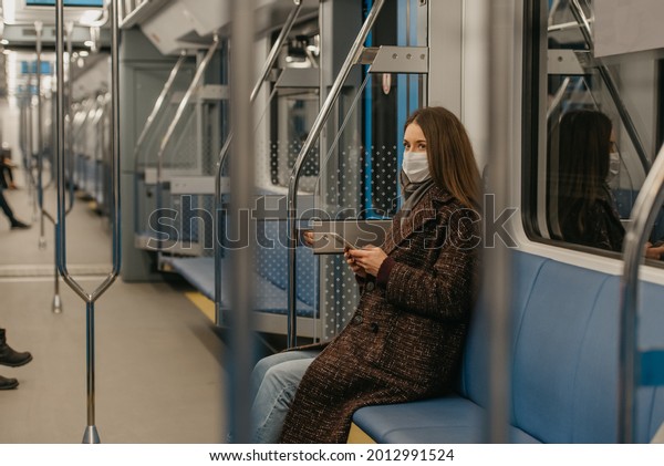 A woman in a medical face mask to avoid the spread\
of coronavirus is sitting holding a smartphone in a modern subway\
car. A girl in a surgical mask against COVID-19 is taking a ride on\
a train.