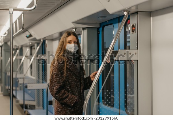 A woman in a medical face mask to avoid the spread\
of coronavirus is standing near doors and staring to the side in a\
modern subway car. A girl in a surgical mask is taking a ride on a\
metro train.