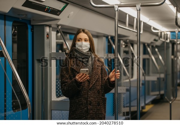 Woman in a medical face mask to avoid the spread\
of coronavirus is standing and holding a smartphone in a modern\
subway car. Girl in a surgical mask against COVID-19 is taking a\
ride on a metro train.
