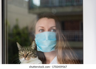 Woman With Medical Face Mask Holding Cat And Sadly Looking Out In The Window Holding Her Cat. Quarantine During Coronavirus Pandemic.