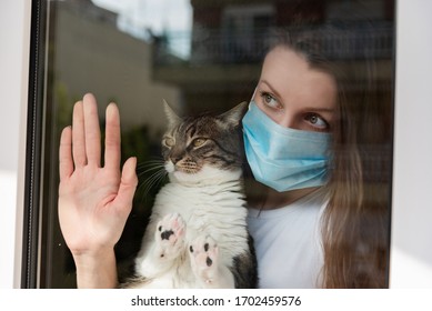 Woman With Medical Face Mask Holding Cat And Sadly Looking Out In The Window Holding Her Cat. Quarantine During Coronavirus Pandemic.