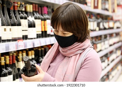 Woman In Medical Face Mask Reading The Label On Red Wine Bottle. Customer In Liquor Store, Concept Of Choosing And Buying Alcohol