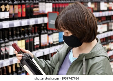 Woman In Medical Face Mask Reading The Label On Red Wine Bottle. Customer In Liquor Store, Concept Of Choosing And Buying Alcohol