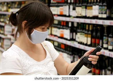 Woman In Medical Face Mask Reading The Label On Red Wine Bottle. Customer In Liquor Store, Concept Of Choosing And Buying Alcohol During Coronavirus Pandemic