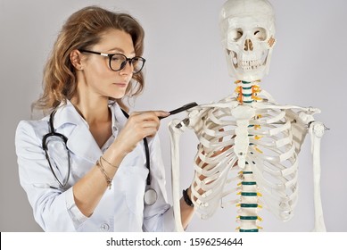 Woman In Medical Gown. Doctor With Skeleton Layout. Girl Medic In Glasses Isolated On A White Background. The Doctor's Assistant Points To The Bone. Study Of Diseases Of The Musculoskeletal System.