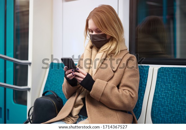 Woman in medical mask with phone in\
her hands sitting in subway car. Coronavirus\
epidemic