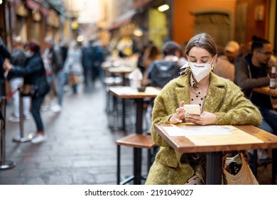 Woman In Medical Mask Sitting With Phone At Outdoor Restaurant On Crowded Street In Bologna City. Concept Of Italian Gastronomic Culture And New Social Rules Related To Pandemic