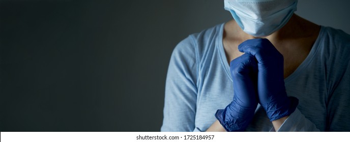 A Woman In A Medical Protective Mask Prays With Faith To GOD. Religion Care During Covid-19 Crisis. Hands In Disposable Gloves Are Folded In Prayer. Online Church Worship. 