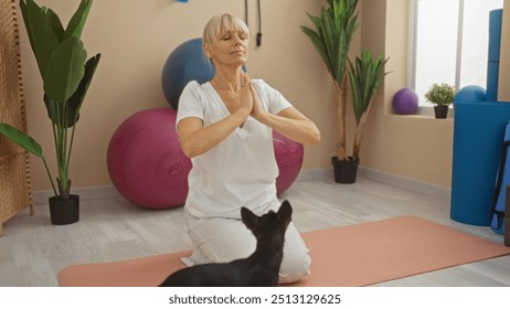 Woman meditating with blonde hair and a black chihuahua dog in a gym filled with exercise balls, plants, and mats - Powered by Shutterstock