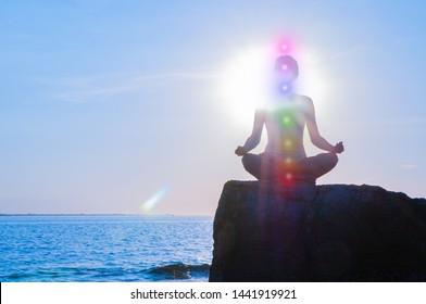 Woman is meditating with glowing seven chakras on stone at sunset. Silhouette of woman is practicing yoga at sunset on the beach. Kundalini meditation.