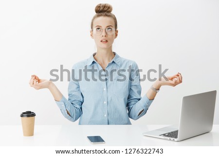 Woman meditating in office sitting near table having break from writing article in laptop, drinking coffee looking up practicing yoga, making nirvana orbs with hands spread aside over gray wall