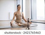 Woman meditating and speaking at camera while sitting in lotus pose. Professional female trainer recording video using tripod and smartphone during physical activity at studio.