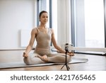 Woman meditating and speaking at camera while sitting in lotus pose. Professional female trainer recording video using tripod and smartphone during physical activity at studio.