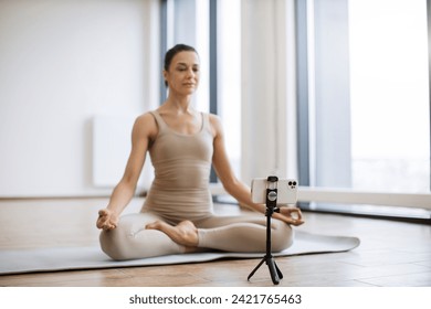 Woman meditating and speaking at camera while sitting in lotus pose. Professional female trainer recording video using tripod and smartphone during physical activity at studio. - Powered by Shutterstock