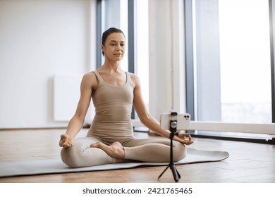 Woman meditating and speaking at camera while sitting in lotus pose. Professional female trainer recording video using tripod and smartphone during physical activity at studio. - Powered by Shutterstock