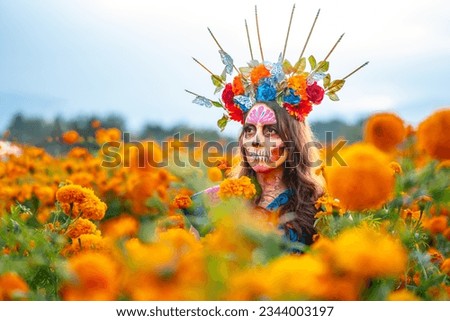 Woman with Mexico Catrina makeup in flower field, serious woman in traditional costume and headwear wearing, standing on field among blooming marigold flowers while looking away. Day of death.