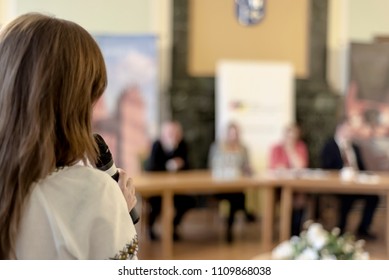 A Woman With A Microphone Performs At A Round Table Meeting. The Photo Is Out Of Focus