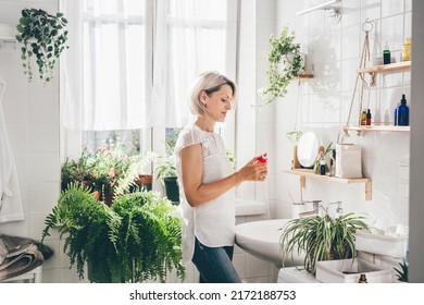 Woman of middle-aged staying in eco friendly white bathroom. Wooden shelves with cosmetics and toiletries against white tile wall. Minimalistic design. Wellness. Comfort home zone - Shutterstock ID 2172188753