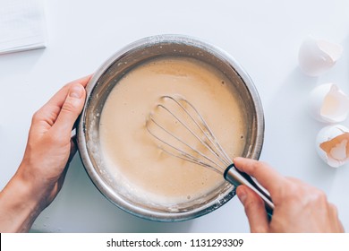 Woman mixing batter, top view. Female's hands holding bowl whisking dough near egg shells on white table.